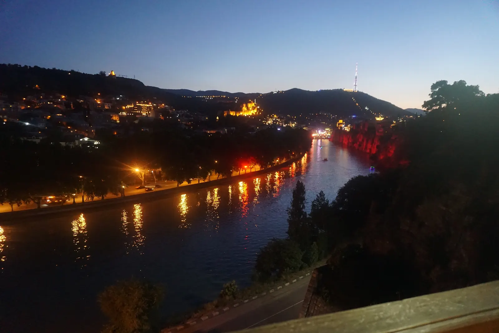 Řeka Kura v Tbilisi za soumraku, s osvětlenými svahy a odlesky městských světel ve vodě